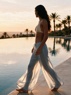 A woman walks barefoot beside an infinity pool at sunset, wearing a white bikini top and flowing white wide-leg pants. Palm trees and a calm pool are visible in the background.