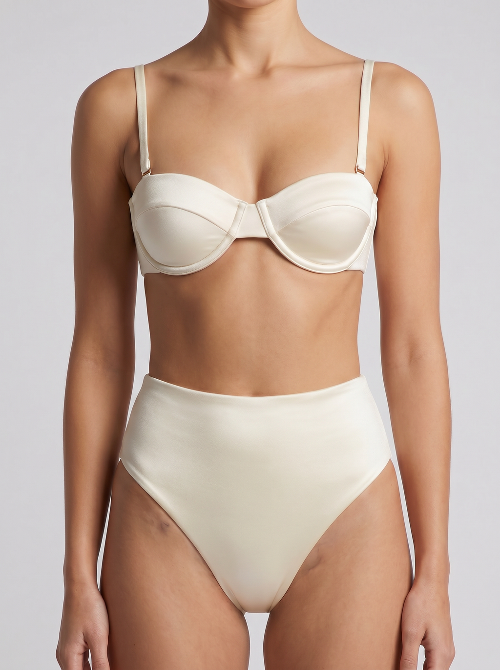 A model wearing a two-piece bikini set in a solid cream color, featuring a structured underwire bra top with thin shoulder straps and high-waisted bottoms, shot against a neutral light gray background.