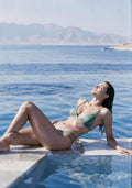 A woman reclining by a swimming pool wearing a sage green string bikini, with blue water and distant mountains in the background.