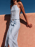A woman wearing a light blue linen two-piece set consisting of a halter crop top and a matching draped wrap skirt, posing against a textured terracotta wall under a clear blue sky.